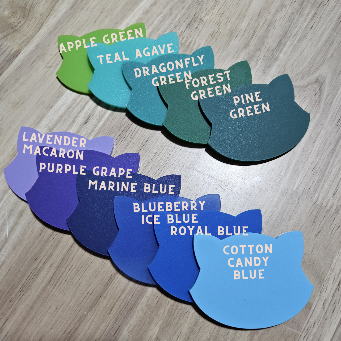 A set of cat-shaped color swatches arranged on a light wood floor, labeled with names like Apple Green, Teal Agave, Dragonfly Green, Forest Green, Pine Green, Lavender Macaron, Purple Grape, Marine Blue, Blueberry Ice Blue, Royal Blue, and Cotton Candy Blue.