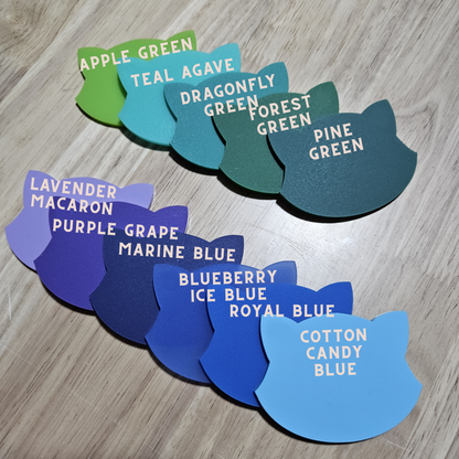 A set of cat-shaped color swatches arranged on a light wood floor, labeled with names like Apple Green, Teal Agave, Dragonfly Green, Forest Green, Pine Green, Lavender Macaron, Purple Grape, Marine Blue, Blueberry Ice Blue, Royal Blue, and Cotton Candy Blue.